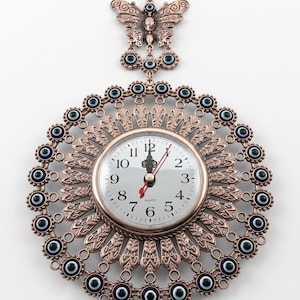 Puede incluir: Un reloj de pared decorativo con un marco de metal color bronce. El reloj tiene una esfera redonda con números y manecillas negras. El marco está adornado con cuentas de ojo turco azules y blancas y una mariposa en la parte superior. El reloj mide unos 25 cm de diámetro.