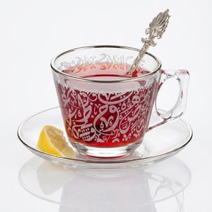 May include: A clear glass teacup with a silver rim and handle, filled with red liquid and a silver spoon. The cup has an etched design. It sits on a matching saucer with a lemon wedge. Tea set.