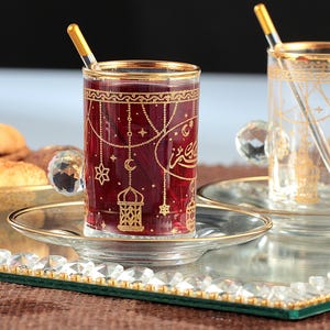 Op de afbeelding: Twee glazen theekopjes met gouden rand en ingewikkelde ontwerpen, waaronder sterren, manen en lantaarns, op een gespiegelde dienblad met een kristallen rand. Een kopje is gevuld met rode thee.