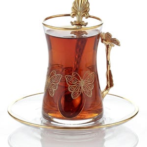 Puede incluir: Una taza de té de vidrio transparente llena de té, con diseños de mariposas doradas y una cuchara a juego. La taza tiene un asa y un borde dorados, y descansa sobre un platillo transparente con borde dorado.