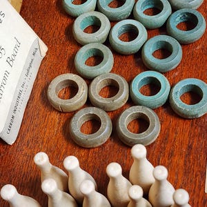 May include: A vintage wooden game set with 10 wooden bowling pins and 15 wooden rings in various colors, including teal, pink, and brown. The rings are stacked in a pyramid shape on a wooden surface. The game set is labeled "Carrom Industries Inc. Ludington, Mich."
