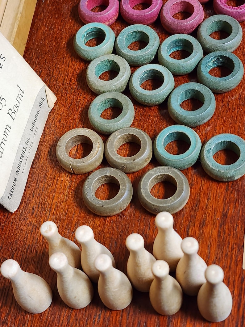 May include: A vintage wooden game set with 10 wooden bowling pins and 15 wooden rings in various colors, including teal, pink, and brown. The rings are stacked in a pyramid shape on a wooden surface. The game set is labeled "Carrom Industries Inc. Ludington, Mich."