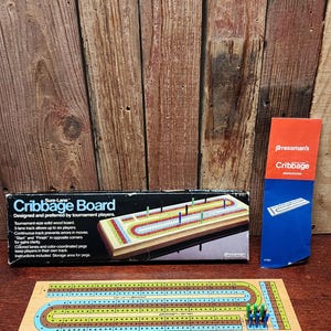 May include: A wooden cribbage board set with a box and instructions. The board has a three-lane track with colored pegs and a storage area. The box reads "Cribbage Board" and "Designed and preferred by tournament players."