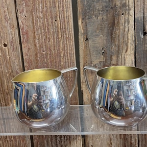 May include: Two silver-colored cream pitchers with gold-colored interiors. Each pitcher has a rounded body, a handle, and a spout. The pitchers are displayed on a clear surface against a wooden background.
