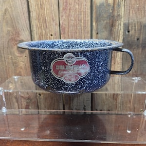 May include: A speckled blue enamelware bowl with a handle. The bowl has a dark blue base with white speckles and a black rim. The bowl has a label that reads "COLUMBIAN Enameled Ware". The bowl is sitting on a clear acrylic shelf.