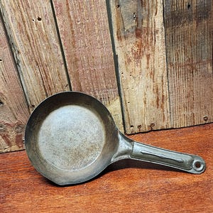 May include: A small, antique metal frying pan with a long handle. The pan is round with a slightly raised edge and a dull, silver finish. The handle has a hole at the end for hanging. The pan rests on a wooden surface.