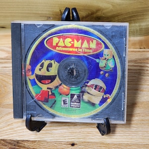 May include: A clear plastic CD jewel case containing the Pac-Man Adventures in Time video game disc. The disc features a yellow Pac-Man character, a mummy, and a knight on a blue background with the title text Pac-Man Adventures in Time.