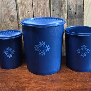 May include: Three navy blue plastic canisters with lids. Each canister has a floral design in a lighter shade of blue. The canisters are cylindrical, with the largest one in the center and two smaller ones on either side.