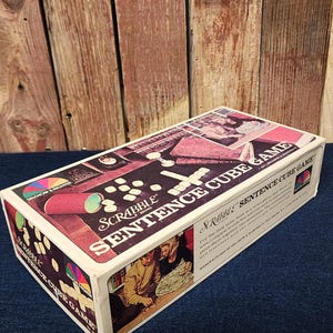 May include: A box for the Scrabble Sentence Cube Game. The box is white with red, blue, and yellow accents. The front of the box features a photo of two people playing the game. The text on the box reads "Scrabble Sentence Cube Game".