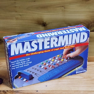 May include: A rectangular blue cardboard box for the Mastermind board game. The front features the title in large white letters, the subtitle The Challenging Game of Logic and Deduction, and an illustration of the game board with colorful pegs.