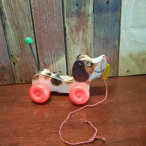May include: A vintage wooden pull-along dog toy with a brown, white, and black spotted body. The toy has two red wheels, a black ear, and a green knob on a stick. A red string is attached to the dog's mouth.