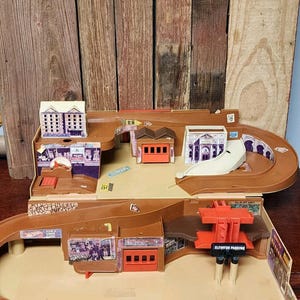 May include: A vintage Hot Wheels playset, featuring a brown plastic design with buildings, a ramp, and a parking structure. The set includes a track for toy cars and various architectural elements.