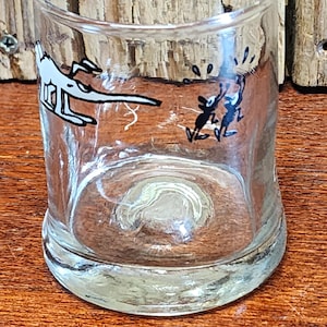 May include: Clear glass shot glass with cartoon illustrations. The glass features a black and white image of a plane and two figures. The glass has a thick base and a slightly curved body.
