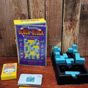 May include: A boxed "Block Block" puzzle game with a yellow and purple box, a deck of cards, and a black tray with turquoise blocks. The box reads "Puzzle Adventures in Three Dimensions!"
