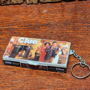 May include: A miniature Clue board game keychain featuring a rectangular plastic case with the classic game box art on the front, depicting various characters in a mansion setting, attached to a silver metal split ring for keys.