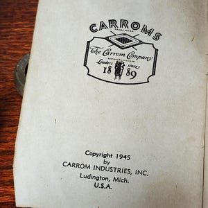 May include: Black and white image of a vintage Carrom Company logo with the text "CARROMS", "The Carrom Company", "Leaders since 1889", and "Copyright 1945 by CARROM INDUSTRIES, INC. Ludington, Mich. U.S.A."