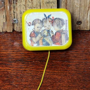 May include: A yellow wind-up music box with a picture of three children on the front. The music box has a pull string and a metal ring.