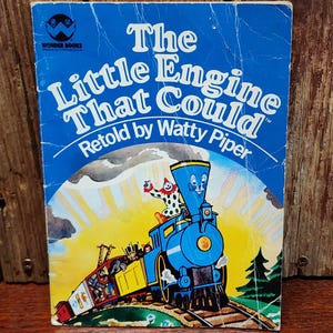 May include: A vintage copy of "The Little Engine That Could" by Watty Piper. The cover is blue with white text and a colorful illustration of a blue train with a smiling face, a clown, and passengers. The book is set against a wooden background.
