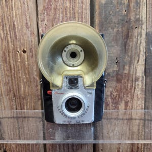 Puede incluir: Cámara vintage Brownie Starflash con una cúpula de flash transparente. La cámara tiene una cara plateada con las palabras "Starflash Camera" y "Brownie" impresas. La cámara es negra y tiene un botón rojo.