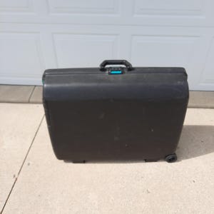 May include: A black rolling suitcase with a top handle and teal accents. The suitcase has a rectangular shape and is sitting on a concrete surface. The wheels are visible on the bottom right side of the suitcase.