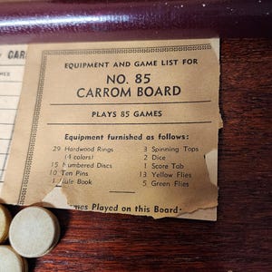 May include: A vintage game board with a list of equipment and games. The board is labeled "No. 85 Carrom Board" and lists 85 games. The equipment includes 29 hardwood rings in 4 colors, 15 numbered discs, 10 ten pins, 1 rule book, 3 spinning tops, 2 dice, 1 score tab, 13 yellow flies, and 5 green flies.