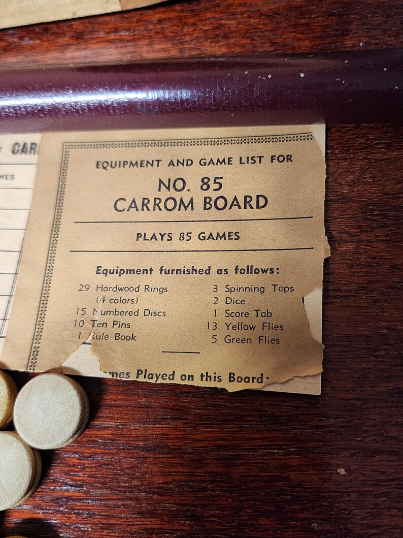 May include: A vintage game board with a list of equipment and games. The board is labeled "No. 85 Carrom Board" and lists 85 games. The equipment includes 29 hardwood rings in 4 colors, 15 numbered discs, 10 ten pins, 1 rule book, 3 spinning tops, 2 dice, 1 score tab, 13 yellow flies, and 5 green flies.