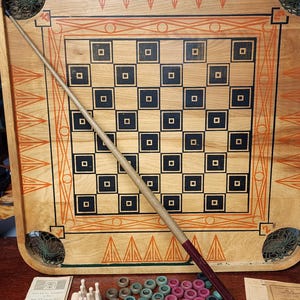 May include: A vintage wooden game board with a checkerboard pattern and a wooden cue stick. The board has a decorative border with orange and black triangles. The game includes wooden pegs, plastic rings, and a scorecard.