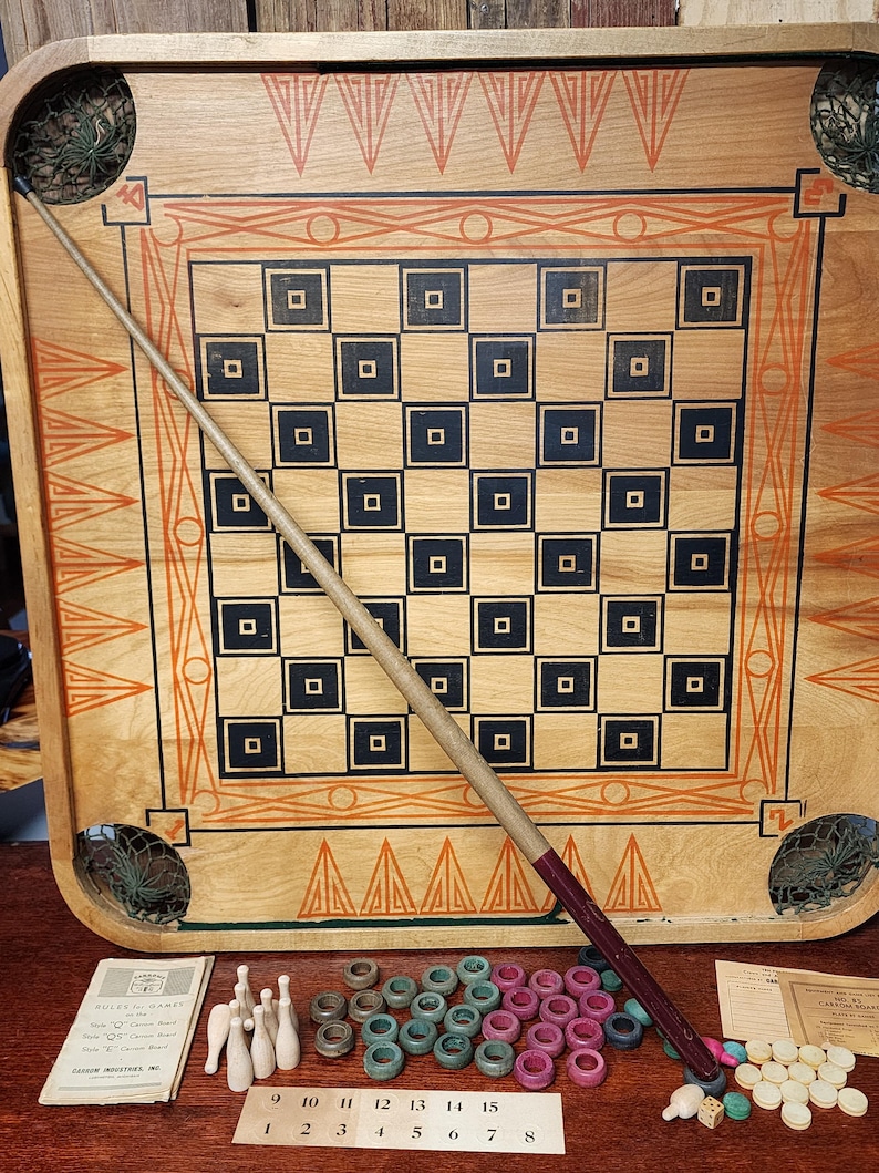 May include: A vintage wooden game board with a checkerboard pattern and a wooden cue stick. The board has a decorative border with orange and black triangles. The game includes wooden pegs, plastic rings, and a scorecard.