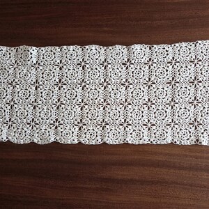May include: A rectangular white crochet table runner featuring a repeating floral motif pattern. The piece is constructed from interconnected circular lace elements with scalloped edges along the length, showcasing a detailed openwork design.