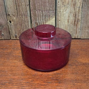 May include: A translucent, ruby-red container with a matching lid. The container is cylindrical with a flat base and a slightly domed lid. A smaller, circular knob sits atop the lid. The container is made of a clear, possibly plastic material.
