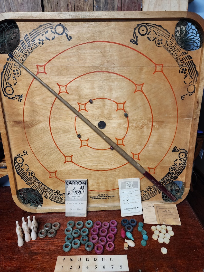 May include: Vintage Carrom board game with wooden pieces, red and black cue stick, and instructions. The board is made of wood and has a circular design with concentric circles. The game is called "Carrom" and is made by "Garcon Industries, Inc."