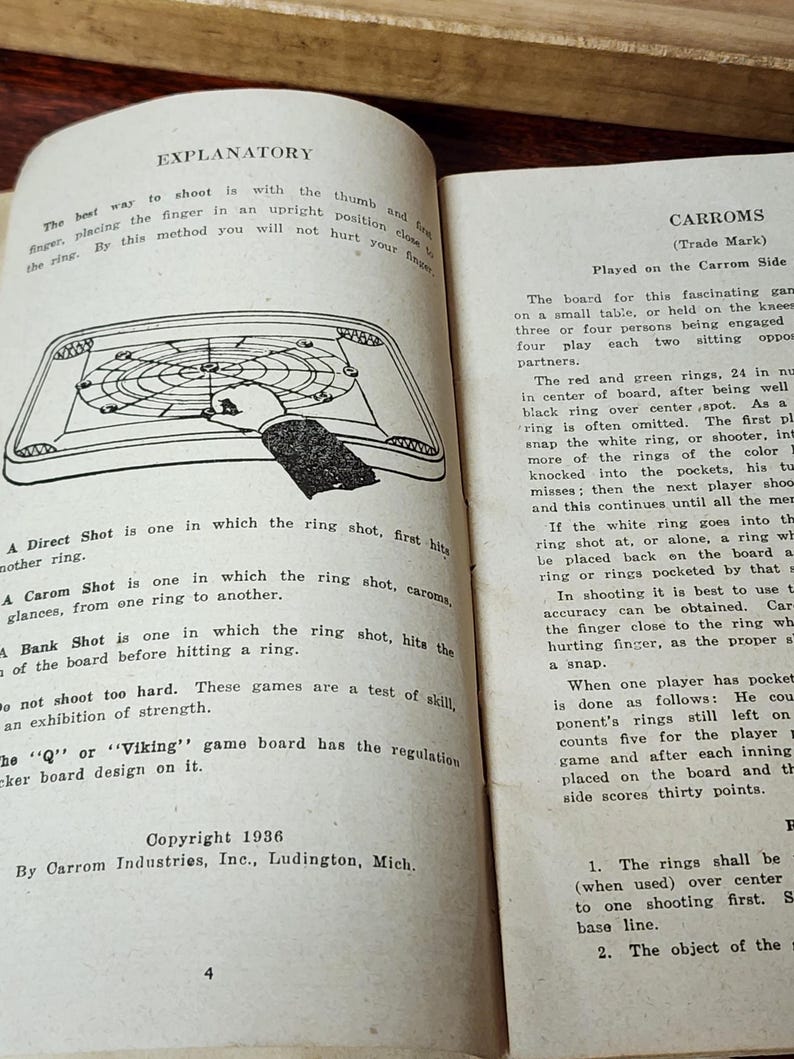 May include: A vintage Carroms game instruction booklet with a black and white illustration of a game board and a hand holding a ring. The text on the page explains the different types of shots in the game, including direct, carom, and bank shots. The booklet is copyrighted 1936 by Carrom Industries, Inc. in Ludington, Michigan.