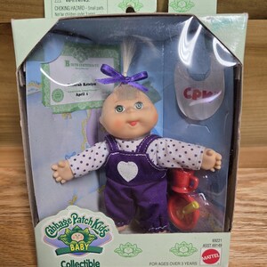 May include: A Cabbage Patch Kids baby doll in original packaging, featuring a light-skinned doll with green eyes and blonde hair in a purple bow. The doll wears white polka-dot long sleeves and purple overalls with a white heart. Includes a birth certificate.