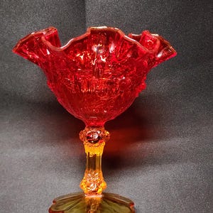 May include: A vibrant red glass candy dish with a ruffled rim. The dish sits on a stem with an amber-colored base and a flower-shaped foot. The glass has a textured, ornate design.