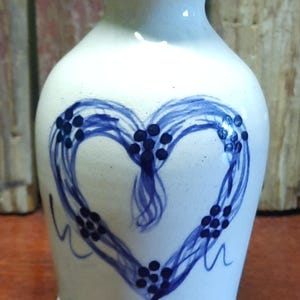 May include: A small, off-white ceramic vase with a blue heart design. The heart is created with flowing lines and dotted accents. The rim of the vase is also trimmed with blue. A decorative piece.