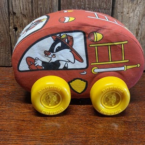 May include: A red and white plush toy fire truck featuring Sylvester the Cat in a firefighter's hat. The toy has yellow wheels with the word "Battat" printed on them. The toy is on a wooden surface.