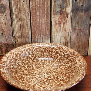 May include: A brown and cream speckled ceramic bowl. The bowl has a wide, shallow shape with a rounded rim. The speckled pattern covers the entire surface of the bowl, creating a rustic, vintage aesthetic. The bowl is likely used for serving or display.