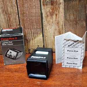 May include: A black Pana-Vue 2 slide viewer with a white top, a box, and a pamphlet. The box is black and silver with the words "Pana-Vue 2" and "Lighted 2x2 Slide Viewer." The pamphlet has instructions in multiple languages.