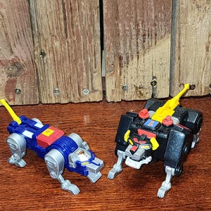 May include: Two robotic animal action figures. One is a blue lion with silver limbs, a yellow tail, and a red square on its back. The other is a black lion with silver limbs, a yellow tail, red accents, and a blue square with the number 1 on its back.