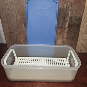 May include: A rectangular, translucent plastic food storage container with a removable white slotted tray insert featuring two side handles. A separate, solid blue rectangular lid with the Tupperware brand name embossed on it is positioned upright.