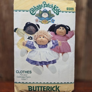 May include: A Butterick sewing pattern envelope for Cabbage Patch Kids clothes, number 6509. The front features three dolls wearing different outfits: a yellow dress with a white apron, a purple dress with a white apron, and a pink polka-dot dress.