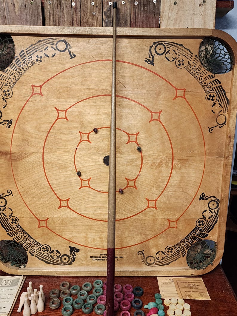 May include: A vintage wooden game board with a circular playing area and a wooden cue stick. The board features a Viking ship design and a net in the corner. The game board is surrounded by a set of colorful wooden rings.