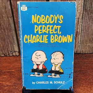 May include: A vintage paperback book titled "NOBODY'S PERFECT, CHARLIE BROWN" with a blue cover and yellow lettering. The cover features cartoon illustrations of Charlie Brown and another character. The book is a Fawcett Crest Book.