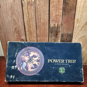 May include: A vintage board game box, navy blue with the title "POWER TRIP" in white text. The box features a circular graphic with zodiac-like figures and the text "for KINETIC INTELLECTS."