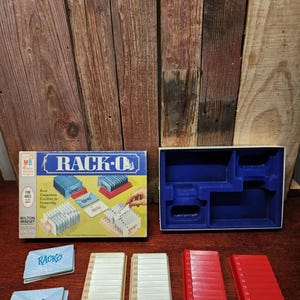 May include: A vintage Rack-O card game set. The box is blue and yellow with the game name in large letters. The set includes cards, card holders, and red and cream-colored racks. The game is designed for ages 8 and up.