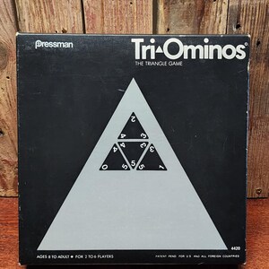 May include: A black Tri-Ominos game box with a white triangle design and the game's name in white. The box features the Pressman logo and text indicating the game is for ages 8 to adult and 2 to 6 players. The game's title is "The Triangle Game."