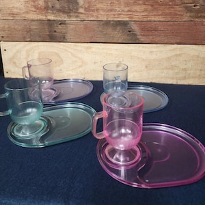 May include: Four sets of colorful, transparent plastic teacups and saucers. The cups and saucers are in shades of pink, blue, and green. The saucers have a unique, curved design. The background is a dark blue surface and a wooden wall.