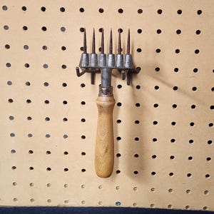 May include: A metal comb with a wooden handle and nine metal teeth. The comb is hanging on a pegboard.