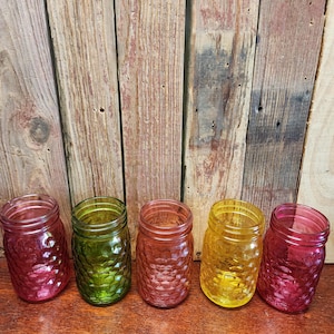 Puede incluir: Cinco tarros de cristal Mason de colores en tonos rojo, verde, naranja y amarillo. Cada tarro tiene un exterior texturizado con diseño de diamante. Los tarros están colocados sobre una superficie de madera con un fondo de madera rústica.