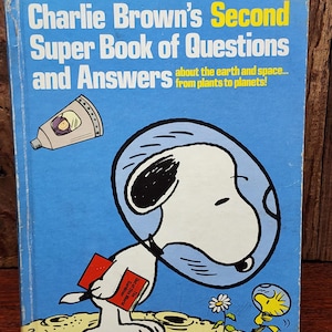 May include: A blue hardcover book titled Charlie Brown's Second Super Book of Questions and Answers about the earth and space... from plants to planets! The cover features Snoopy and Woodstock in space helmets on the moon, with a space capsule.